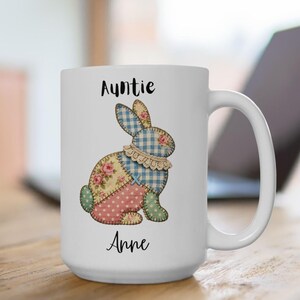 May include: White ceramic mug with a patchwork bunny design. The bunny features blue gingham, pink polka dots, and floral patterns. The mug is inscribed with the words "Auntie Anne" in black script.