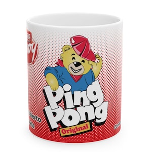 May include: White ceramic mug featuring a cartoon bear wearing a red cap and blue shirt. The mug has the words "Ping Pong Original" in large letters, set against a red and white dotted background. The Nestle logo is also visible.