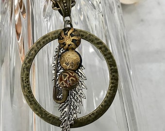 May include: A gold necklace with a large, hammered circle pendant. The pendant has a silver feather charm and three gold and black beads hanging from it.