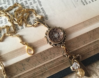 May include: A gold chain necklace with a bronze pendant featuring a star design. The pendant is attached to a gold chain with a small charm and a crystal bead.