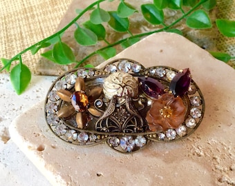 May include: A vintage-style brooch with a gold-tone metal base and a variety of colorful glass stones. The brooch features a flower design with a brown center, two purple stones, and a white and gold swirl design.