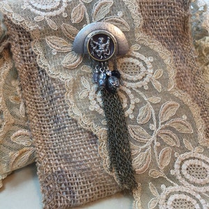 May include: A silver and black pendant with a chain and a small figure of a person in the center. The pendant is on a burlap and lace background.
