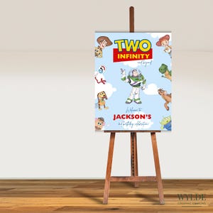 Puede incluir: Un letrero azul claro sobre un caballete de madera con personajes de la película Toy Story. El letrero dice "TWO INFINITY and beyond" con el texto "Welcome to JACKSON'S 2nd birthday celebrations!" El letrero tiene ilustraciones de Woody, Buzz Lightyear y otros personajes.