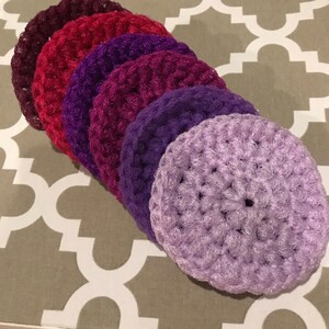 May include: Six crocheted scrubbers in shades of purple, red, and pink. The scrubbers are round and have a textured surface.