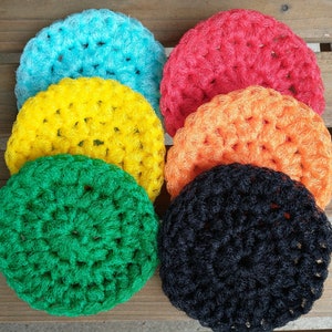 May include: A collection of six crocheted circular items in various colors: turquoise, red, yellow, orange, green, and black. The items are arranged on a wooden surface, showcasing their handmade texture and vibrant hues. These could be used as coasters or decorative elements.