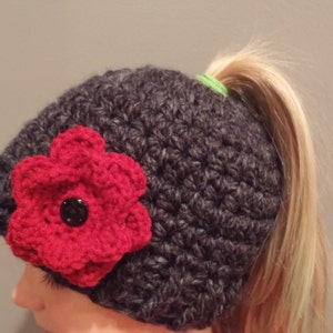 Può includere: Un cappello a uncinetto grigio scuro con un fiore a uncinetto rosso e un elastico per capelli verde. Il cappello è progettato per essere indossato con una coda di cavallo.