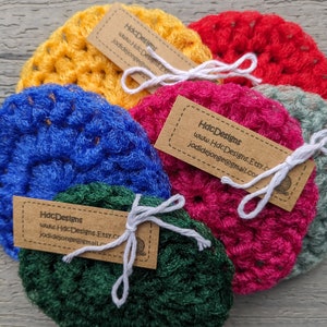 May include: Five crocheted scrubbers in different colors, including yellow, blue, pink, green, and gray. Each scrubber has a small brown tag attached with the text "HdcDesigns www.HdcDesigns.Etsy.com jodidejonge@gmail.com".
