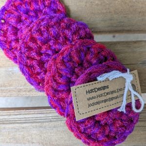 May include: Four handmade crochet coasters in shades of purple and red. The coasters are round and have a textured appearance. A small tag with the text "HdcDesigns" is attached to the coasters.