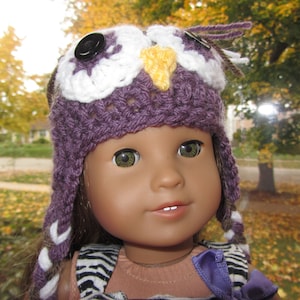 May include: A doll wearing a purple and white crocheted owl hat with a yellow beak and black button eyes. The doll has brown hair and is wearing a striped shirt.