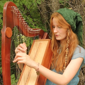 May include: A woman plays a wooden harp with a green knitted hat on her head. She is wearing a blue shirt and has long red hair with a brown feather braid.