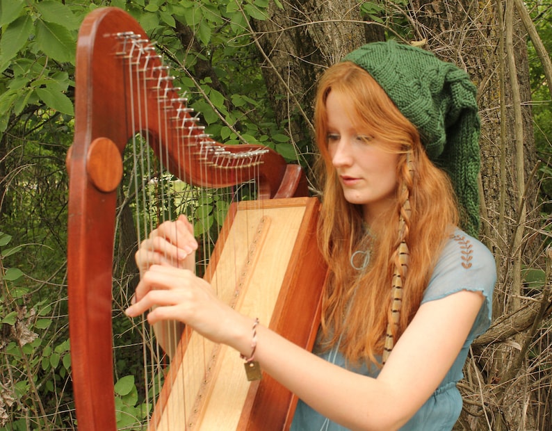 May include: A woman plays a wooden harp with a green knitted hat on her head. She is wearing a blue shirt and has long red hair with a brown feather braid.