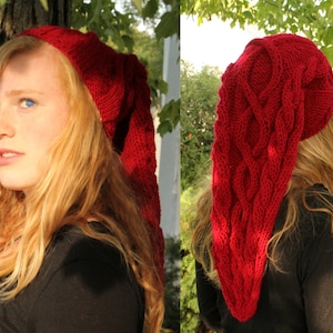 May include: A red knitted hat with a long, pointed tail. The hat features a cable knit pattern and is being worn by a person with long, red hair.