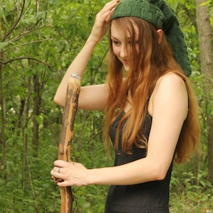 May include: A woman wearing a green knitted hat with a long pointed tip, a black dress, and a wooden walking stick. She is standing in a woodland setting.