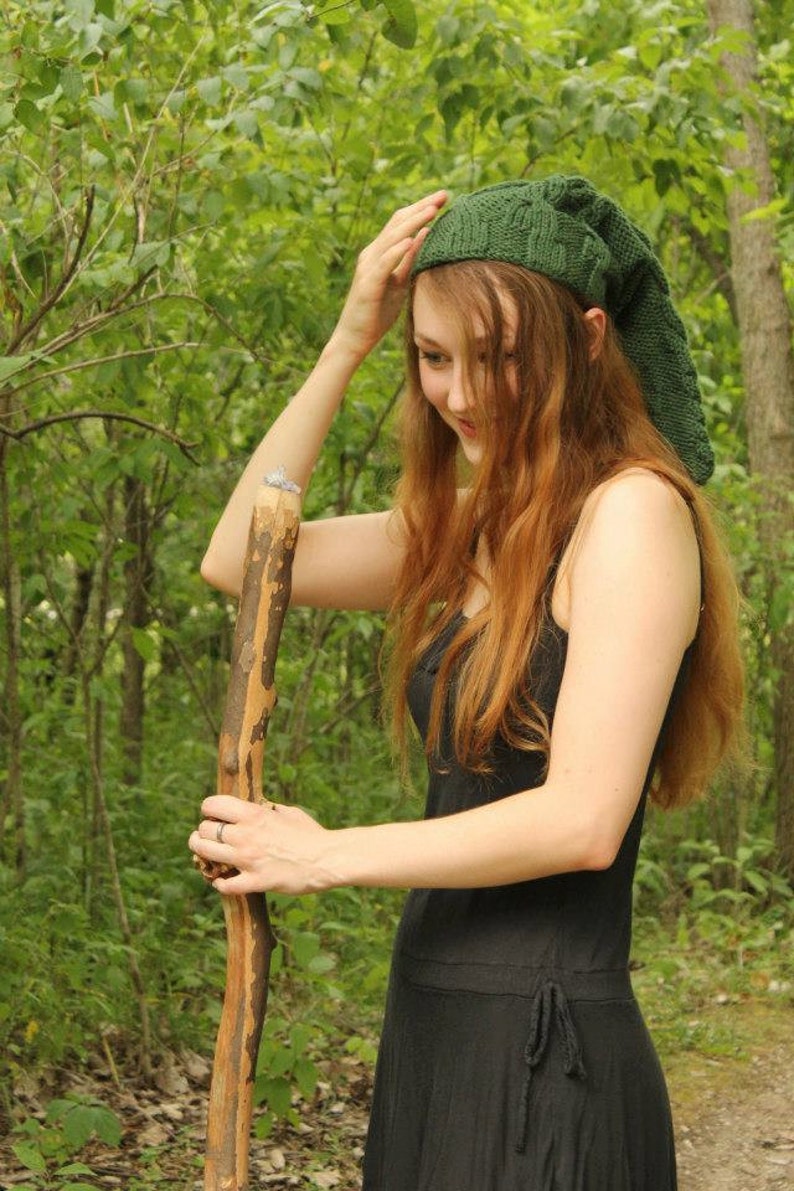May include: A woman wearing a green knitted hat with a long pointed tip, a black dress, and a wooden walking stick. She is standing in a woodland setting.