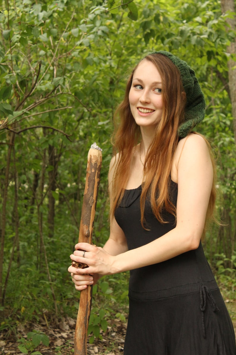 May include: A woman with long red hair wearing a green knitted hat and a black dress holds a wooden walking stick.