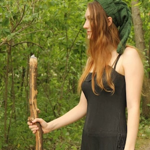 May include: A woman wearing a green knitted hat with a long pointed tip, a black sleeveless dress, and holding a wooden walking stick. She is standing in a woodland setting.