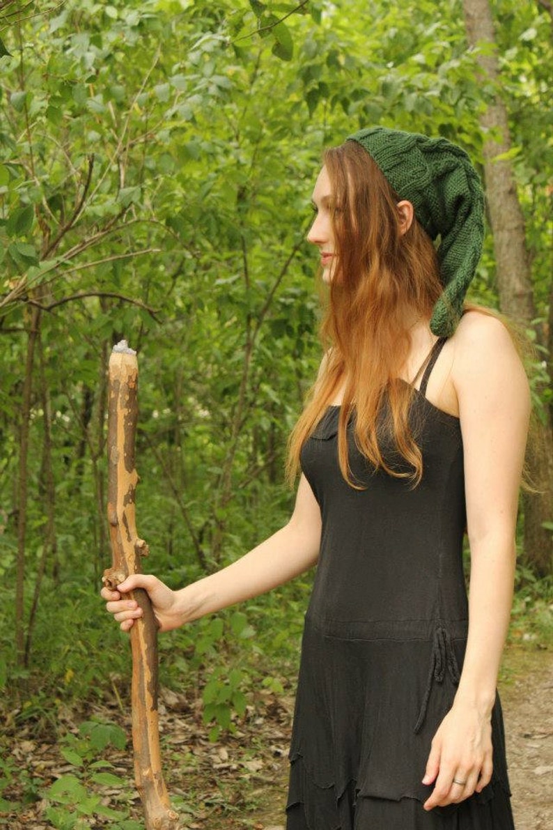 May include: A woman wearing a green knitted hat with a long pointed tip, a black sleeveless dress, and holding a wooden walking stick. She is standing in a woodland setting.
