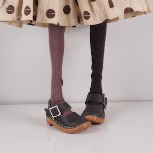 May include: A doll's legs and feet are visible, wearing mismatched socks and brown leather shoes. The doll's skirt is beige with brown polka dots. The shoes have a buckle closure and a wooden sole.