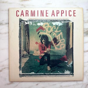Puede incluir: Portada de álbum vintage con el texto "CARMINE APPICE" en rojo. La ilustración muestra a un hombre con chaqueta roja y gafas de sol arrodillado frente a un grafiti que dice "ROCK ERS". La portada tiene un aspecto envejecido y desgastado.