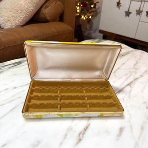 Puede incluir: Joyero abierto con un estampado floral en amarillo, verde y blanco. El interior está forrado con satén melocotón claro y un material dorado similar al terciopelo con múltiples ranuras para anillos y otras joyas. La caja tiene un borde dorado.