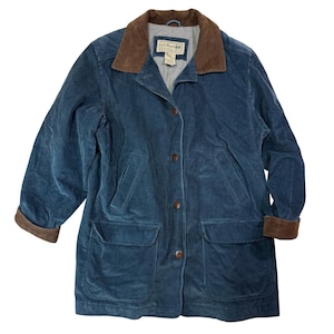 Op de afbeelding: Een blauwe corduroy jas met een bruine corduroy kraag en manchetten. De jas heeft een knoopsluiting, twee voorzakken en een klein label op de borst. De jas is middellang.