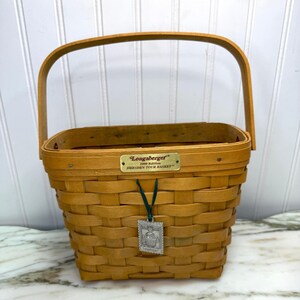 May include: A woven rectangular basket with a handle. The basket is made of light brown wood and features a metal tag that reads "Longaberger 1998 Edition Dresden Tour Basket". A small charm hangs from a green ribbon.