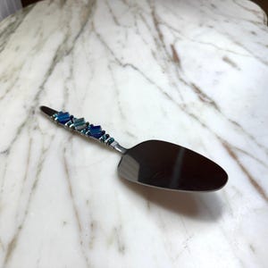 Puede incluir: Una espátula para pastel de acero inoxidable con un mango decorativo. El mango está adornado con cuentas azules y turquesas y alambre. La espátula está sobre una superficie de mármol blanco y gris.