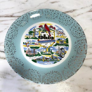 Könnte beinhalten: Eine Vintage-Souvenirplatte mit hellblauem Rand und einem zentralen Design mit Wahrzeichen von Ohio. Die Platte hat goldene Akzente und den Text "Ohio Mother of Presidents". Das zentrale Design zeigt einen roten Kardinal.