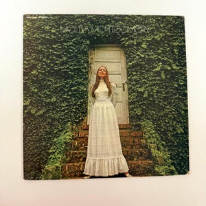 Puede incluir: Portada del álbum con Melba Montgomery con un vestido blanco frente a una puerta. La puerta está rodeada de hiedra verde. El título del álbum "Melba Montgomery" está impreso en la parte superior.