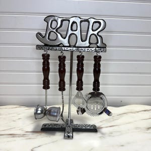 May include: A silver-toned bar tool set with a "BAR" sign. The set includes a jigger, bar spoon, bottle opener, and strainer, each with dark brown wooden handles. The tools hang from a decorative metal stand.