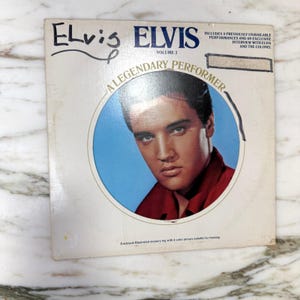 May include: Vintage Elvis Presley "A Legendary Performer" Volume 3 album cover. The cover shows a portrait of Elvis in a red shirt against a blue backdrop. The album title and artist's name are printed in blue and black.
