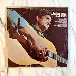 May include: Vintage vinyl record album cover featuring Johnny Cash playing a guitar. The album cover has a sepia tone with the artist's name in white lettering. The album's song titles are listed on the right side of the cover.