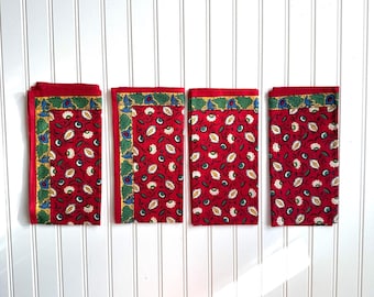 Op de afbeelding: Vier gevouwen rode stoffen servetten met een bloemenpatroon. Elke servet heeft een rode rand en een decoratieve rand met groene, blauwe en gele accenten. De servetten zijn gerangschikt op een wit oppervlak.