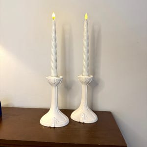 May include: Two white ceramic candle holders with lit, twisted candles. The candle holders have a detailed base and a slender stem. The candles have a realistic flame effect, creating a warm ambiance. The overall design is elegant and suitable for home decor.