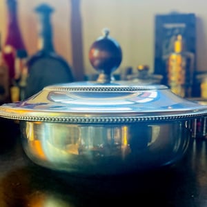 Puede incluir: Un plato de servicio plateado pulido con tapa redondeada y un pomo esférico de color marrón oscuro. La tapa presenta un detalle decorativo de cuentas en el borde y el cuenco tiene un acabado metálico liso y reflectante.