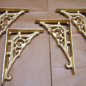 May include: Four ornate, gold-colored shelf brackets. Each bracket has a decorative, openwork design and a curved hook at the bottom. The brackets are made of metal and are likely for supporting shelves or other items.