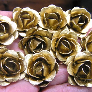 May include: A collection of ten gold-colored metal rose flower embellishments. The roses are arranged in a hand, with the petals of each rose overlapping.