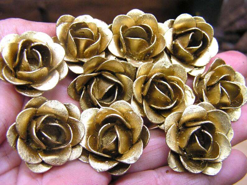 May include: A collection of ten gold-colored metal rose flower embellishments. The roses are arranged in a hand, with the petals of each rose overlapping.