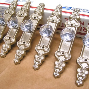 May include: Six ornate gold-toned cabinet door pulls with clear glass crystal knobs. Each pull has a decorative design with a floral motif.