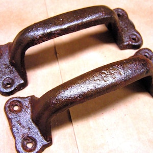 May include: Two antique, rusty brown cast iron handles. Each handle has a curved grip and two mounting plates with holes for screws. The handles have a weathered appearance, suggesting age and use.