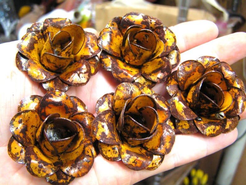 Puede incluir: Cinco adornos de metal con forma de rosa, cada uno con un acabado amarillo brillante y manchas de color marr&oacute;n oscuro que parecen &oacute;xido en los p&eacute;talos superpuestos. Estas piezas florales decorativas tienen una estructura redondeada y multicapa.