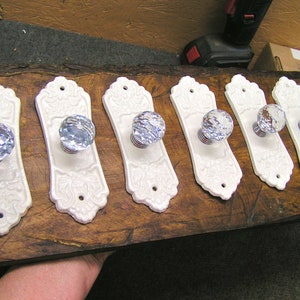May include: Six white ceramic door knobs with ornate designs and clear crystal glass knobs. The knobs are arranged in a row on a wooden surface.
