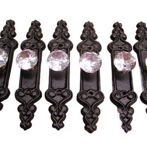 May include: A set of six drawer pulls, each featuring a long, black, ornate metal backplate with intricate scrollwork and a single, clear, faceted glass knob attached to the center. The hardware is arranged in a vertical row on a white background.