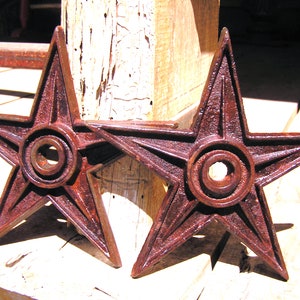 May include: Two rusty, red-brown cast iron star-shaped decorative pieces with a circular center hole. The stars are attached to a wooden surface.