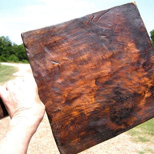 May include: A square wooden cutting board with a dark, stained finish and a visible, textured wood grain. The board features a thick handle on one side with a small hole containing a thin leather cord for hanging, showcasing a rustic appearance.