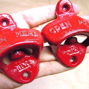 May include: Two red metal bottle openers with the words "Open Here" on them. The openers are mounted on a wall.
