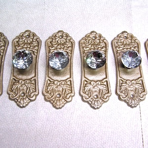 May include: A set of six rectangular gold-toned metal drawer pulls, each featuring an ornate embossed floral pattern at the top and bottom, centered with a clear, multifaceted, circular glass knob that reflects light with a brilliant sparkle.