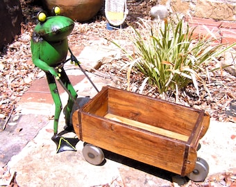 Può includere: Una scultura di rana in metallo verde con occhi gialli e piedi palmati neri, in piedi mentre tiene il manico di un carro rettangolare in legno. Il carro presenta una finitura in legno naturale, accenti in metallo scuro e quattro ruote metalliche.