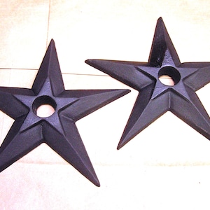May include: Two black, star-shaped decorative objects. Each star has five points and a circular hole in the center. The objects have a textured surface and are likely made of cast iron or a similar material.