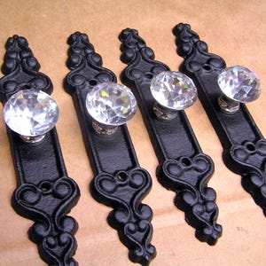 May include: Four drawer pulls featuring clear, multifaceted glass knobs attached to elongated, matte black metal backplates. Each backplate displays a decorative, scroll-like design with two circular screw holes for mounting purposes.